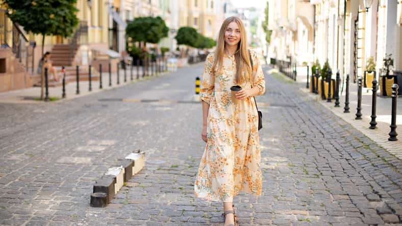 smiling caucasian woman blonde in dress with coffee takeaway walks on the cobblestones against the backdrop of the street and green trees in summer