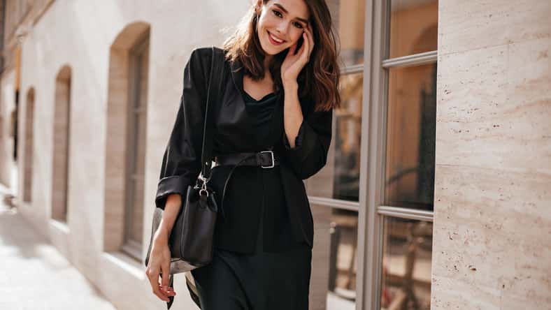 Cute slim girl with brown wavy hairstyle, bright makeup, dark outfit of long dress, oversized jacket, belt on waist and trendy bag, standing on daylight street in city and smiling