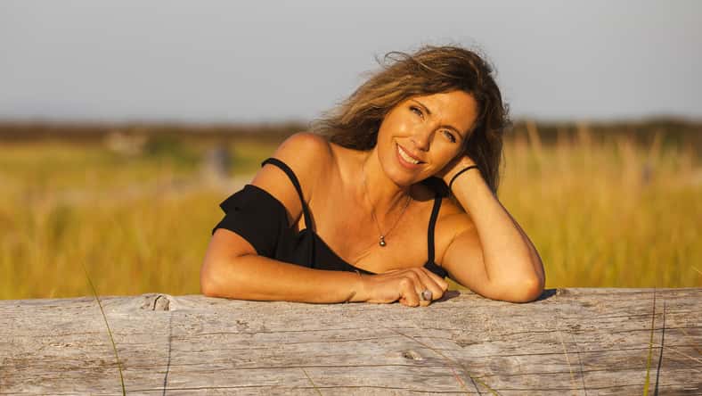 Pretty middle aged woman smiles while leaning on driftwood log outside in nature during the golden hour