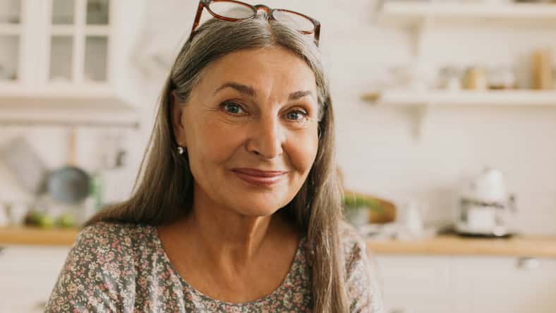 Pretty grey-haired elderly woman keeping glasses on forehead posing in front of camera with adorable smile, wearing flowered dress, sitting against modern bright kitchen room. Lifestyle concept