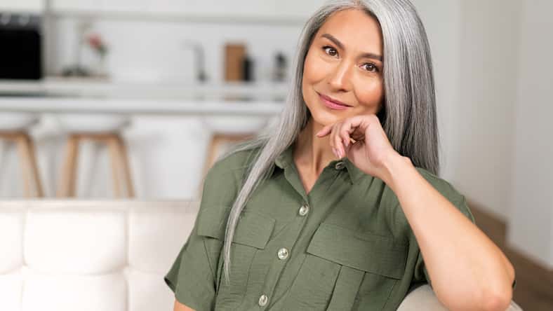 Close-up portrait of charming middle-aged woman with long silver hair sitting in relaxed athmosphere, mature 50s asian female resting at home, looking at camera and smiling with calm smile