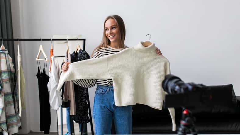 Fashion blogger woman in jeans and turtleneck showing casual colorful shirts on camera. Stylist influencer girl showing trendy clothes filming vlog episode for her channel. Opinion leader sets trends.