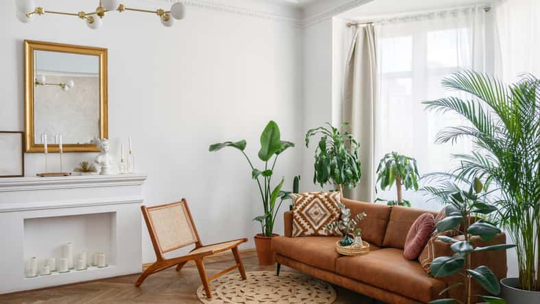 Apartment with bright living room in boho style. Interior design with brown couch, new armchair, decorative fireplace with candles, green house plants in clay pots and rug on the wooden floor