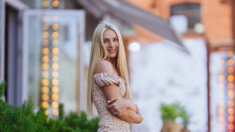 Gorgeous Blonde young woman on weekend waking outside looks at camera toothy smiling against blurry food court with plants. Stunning Swedish girl in love waiting for boy friend on Valentines day.