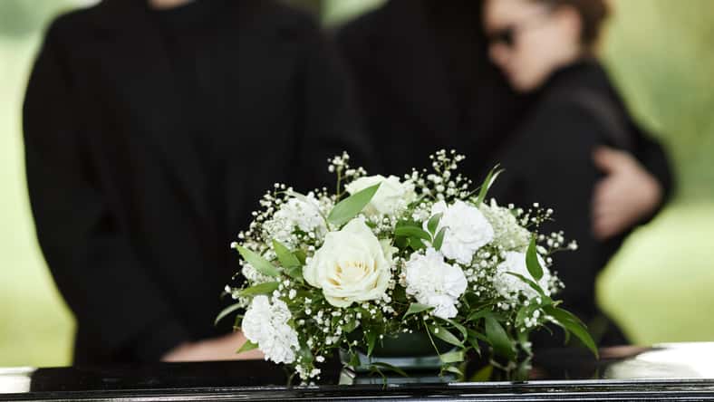 Close up of flower bouquet on coffin with people wearing black in background, outdoor funeral ceremony, copy space