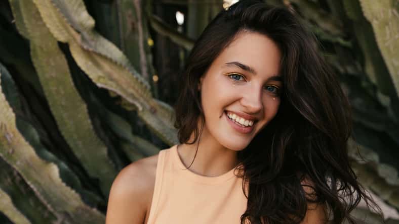 Close-up of positive young caucasian woman with stylish make-up looking at camera and smiles. Brunette wears peack color top. Concept of real positive emotions.
