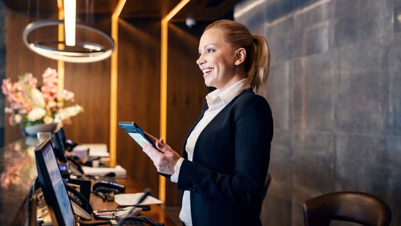 A friendly receptionist is talking to a client while making a reservation on a tablet at hotel reception.