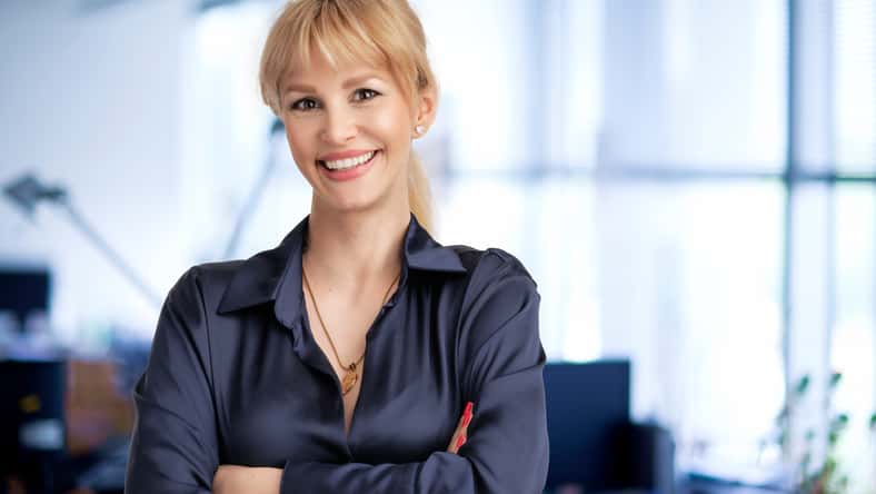 Happy business woman with blond hair standing at the office with folded arms.