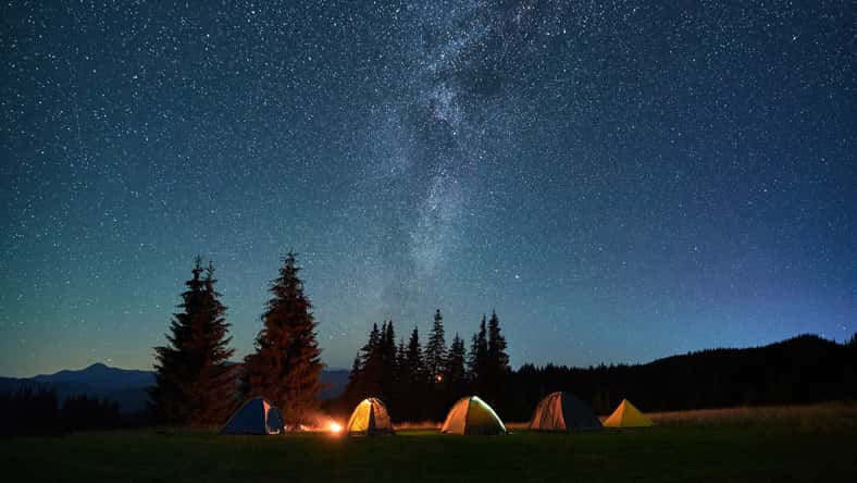 Night camping in mountains under starry sky. Tourist tents in campsite near burning campfire under beautiful sky full of stars with Milky way above forest. Concept of tourism and traveling.