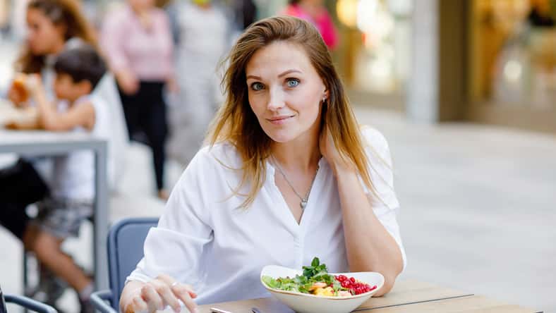 Young beautiful woman eating fresh salad in outdoor restaurant. Woman enjoying lunch bowl with fresh vegetables. Healthy food. On summer day