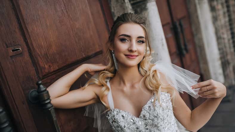 Beautiful young smiling bride curly blonde in white lace long dress stands, walks against the background of an old retro church, wooden doors. Wedding photography, portrait.