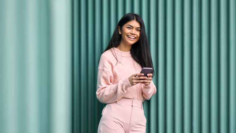 Feliz chica adolescente asiática con atuendo rosa usando aplicaciones de teléfonos móviles al aire libre mirando la cámara. Modelo femenino y sonriente positivo que sostiene el teléfono inteligente riendo de pie con fondo de pared azul.
