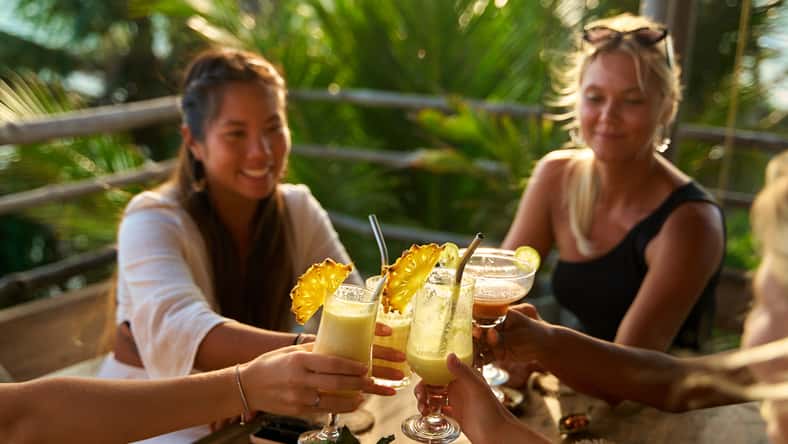 Diverse group of women at tropical restaurant toast with cocktails. Celebration. Vacation mood. Fun bachelorette party, on travel. Laughter in paradise. Enjoying sun vibes together. Happy moments.