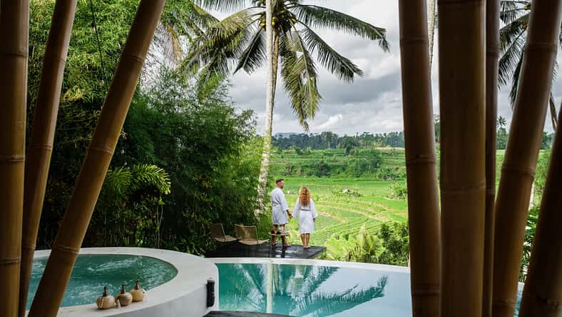Romantic Couple in Bathrobes Enjoying Scenic Tropical Landscape from Luxury Infinity Pool Overlooking Rice Fields and Palm Trees in Bali, Indonesia.