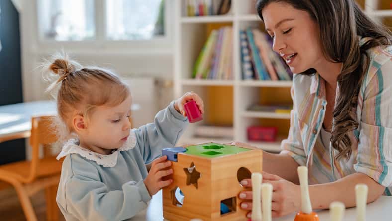 A young woman and a little girl play with educational toys at a preschool or daycare, engaging in learning activities with a shape sorter in a bright, cheerful classroom