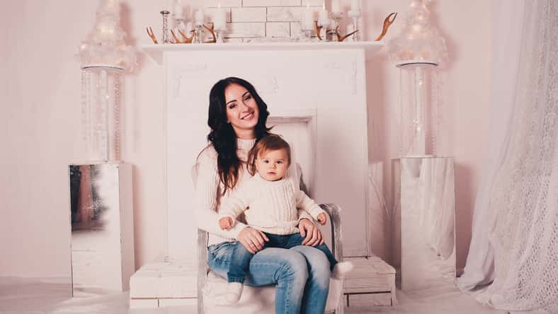 Smiling mother sitting with baby girl in armchair at home. Looking at camera. Motherhood.