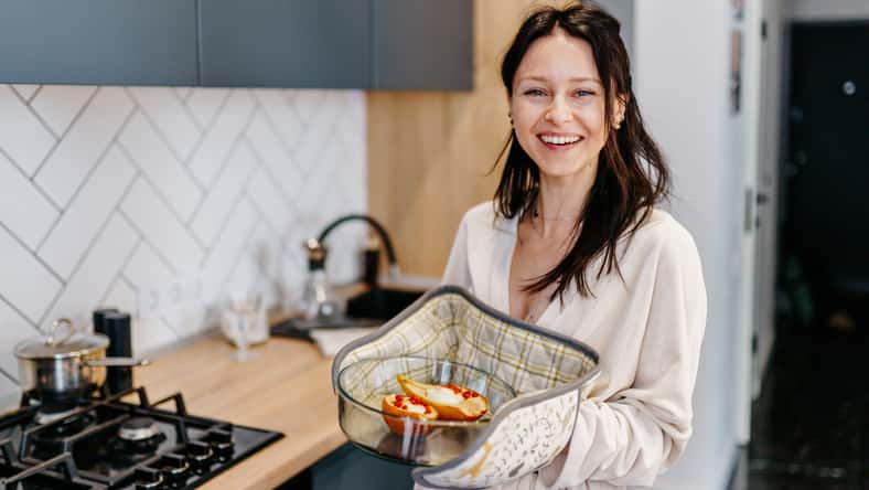 Mother in her 40s smiling and holding baked pears in glass dish, standing in modern home kitchen. Joyful moment after home cooking. Concept of healthy lifestyle, positive motherhood, simple pleasures.