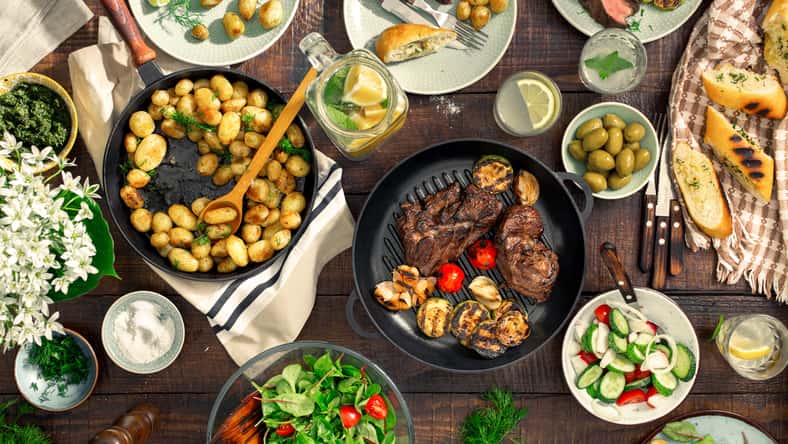 Dinner table with meat grill, roast new potatoes, vegetables, salads, sauces, snacks and lemonade, top view