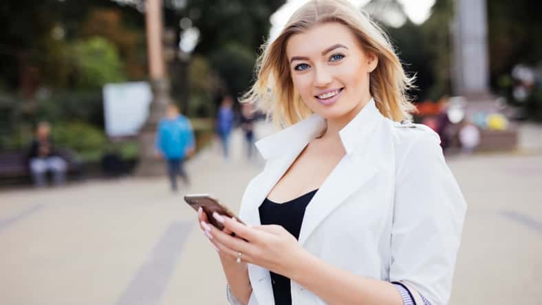gorgeous beautiful young woman with blonde hair messaging on the smart-phone at the city street background. pretty girl having smart phone conversation in sun flare.