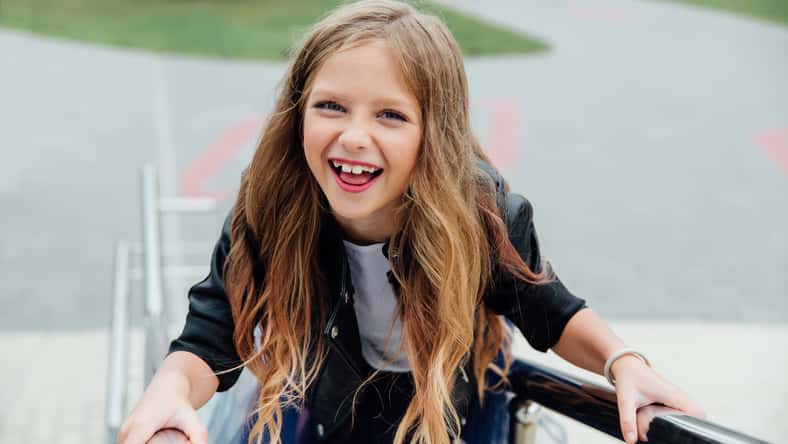 Urban fashion portrait of a teenage girl in the street at the railing on the stairs. Long hair, blue skirt, black leather jacket. Carefree childhood. Emotions, grimaces, a smile.