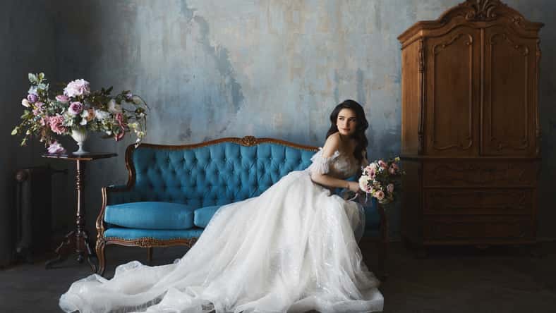 beautiful brunette model girl in stylish and fashionable lace wedding dress with naked shoulders sits on the antique sofa and posing in luxury vintage interior.
