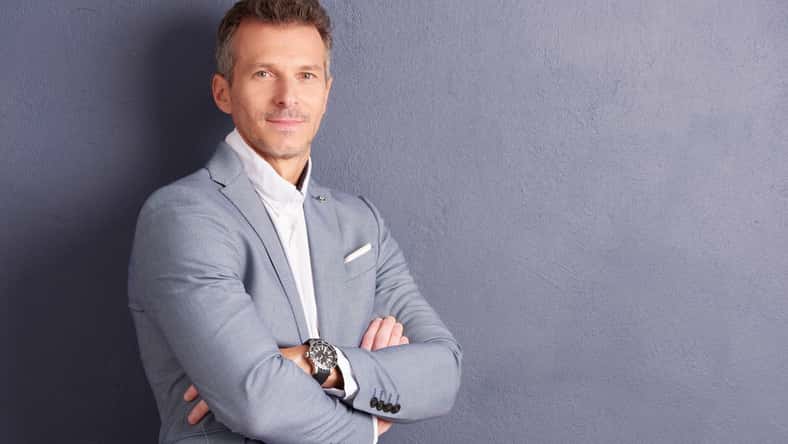 Portrait shot of confident middle aged businessman with folded arms standing at grey wall and looking at camera.