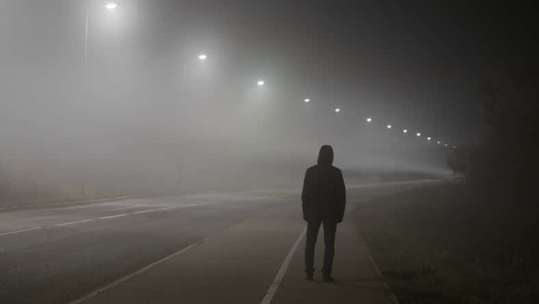 Young man alone slowly walking under white street lights in night. Dark time. Peaceful atmosphere in mist. Foggy air. Back view.
