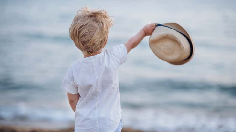 A rear view of small blond toddler boy with hat standing on beach on summer holiday.