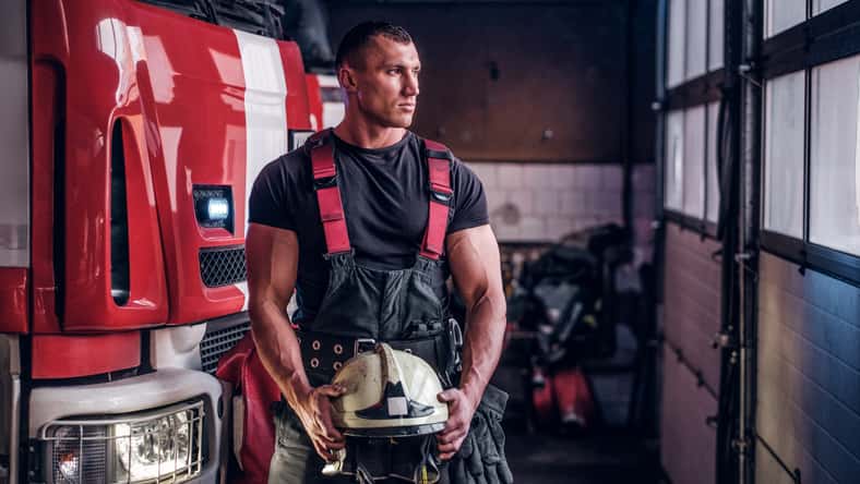 Muscular fireman holding a protective helmet in a garage of a fire department, standing next to a fire engine and looking outside