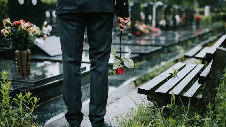 Elegant sad elderly man with black suite and umbrella walking alone on graveyard. Rainy day.