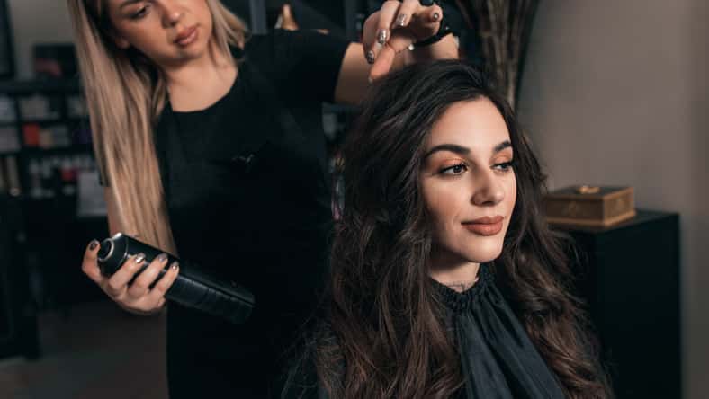 Female hairdresser makes hairstyle on young woman with brunette hair in salon.