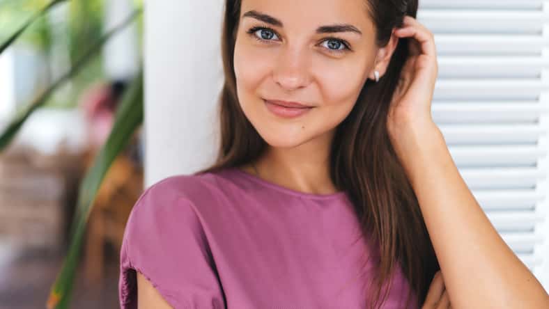 Close-up portrait of beautiful young brunette woman with amazing green eyes in modern white style cafe. Girl in pink dress posing near bright wooden wall, she has natural make-up