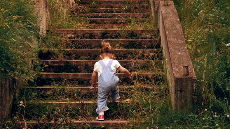 A little girl, 2 years old, with a ponytail and in a jumpsuit, climbing up the steps of the old stone, overgrown grass stairs. Concept of overcome. Cottagecore aesthetics concept.