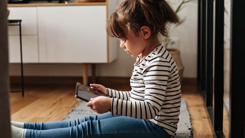 Portrait of little girl in jeans and t-shirt sitting on carpet and holding tablet