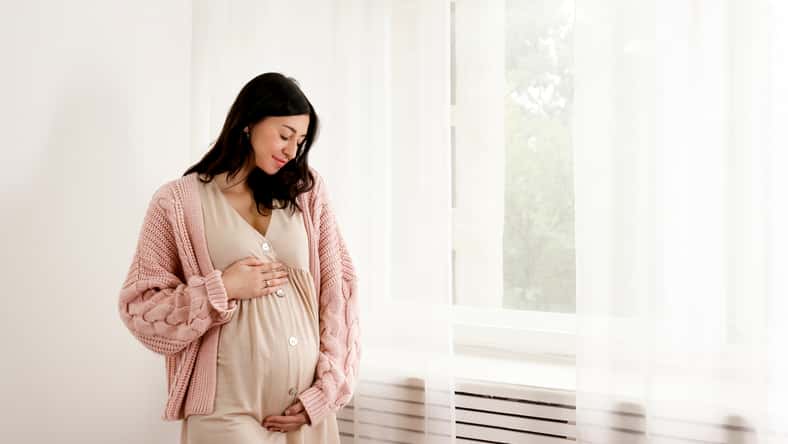 Young beautiful woman on second trimester of pregnancy. Close up of pregnant female in pink oversized knitted sweater with arms on her round belly. Expecting a child concept. Background, copy space.