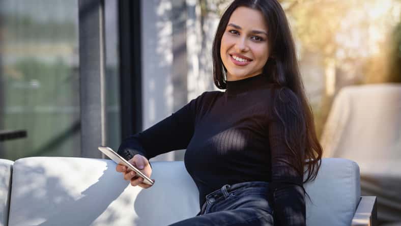 Portrait of a young woman with long, black hair sitting on a terrace and holding her mobile phone
