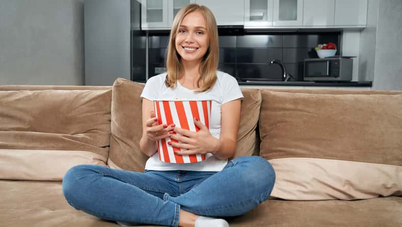 Happy woman in casual outfit sitting with crossed legs on couch with big striped bucket of popcorn. Smiling blonde relaxing at home and watching movie.
