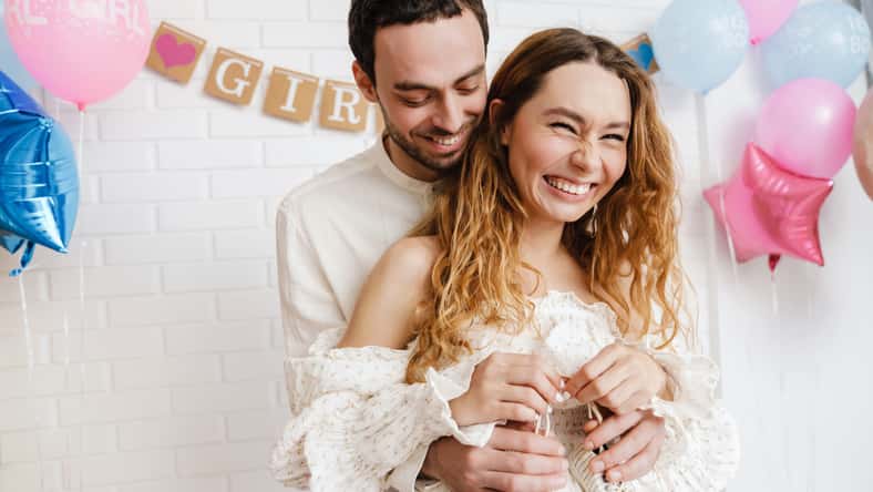 Young happy couple posing with baby shoes during gender reveal party indoors