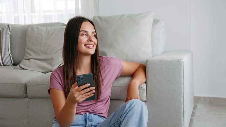 Portrait of young brunette woman wearing mom jeans sitting on the floor leaning on the couch texting and smiling. Joyful female having fun video phone call. Background, copy space, close up.