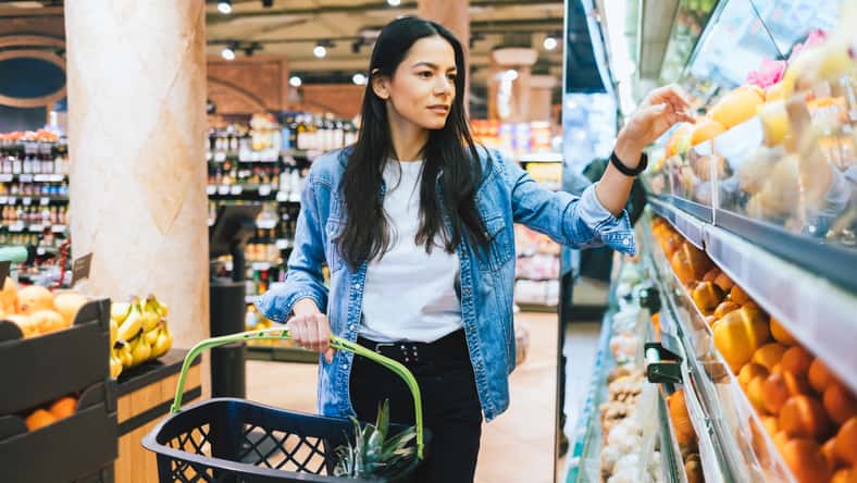 Young woman doing grocery shopping in supermarket, choosing citruses near shelf with fresh organic fruits and vegetables