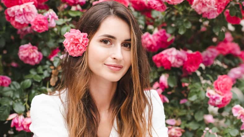 Smiling beautiful woman with long blonde hair and rose flower over blooming nature background outdoor. Attractive female with healthy skin and floral hairstyle. Springtime.