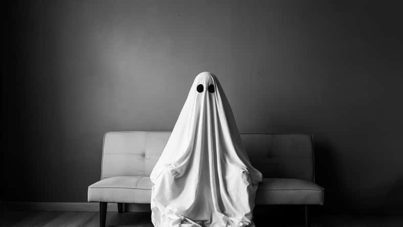 Ghost covered with a white ghost sheet on a sofa in the living room. Halloween concept. Black and white photo