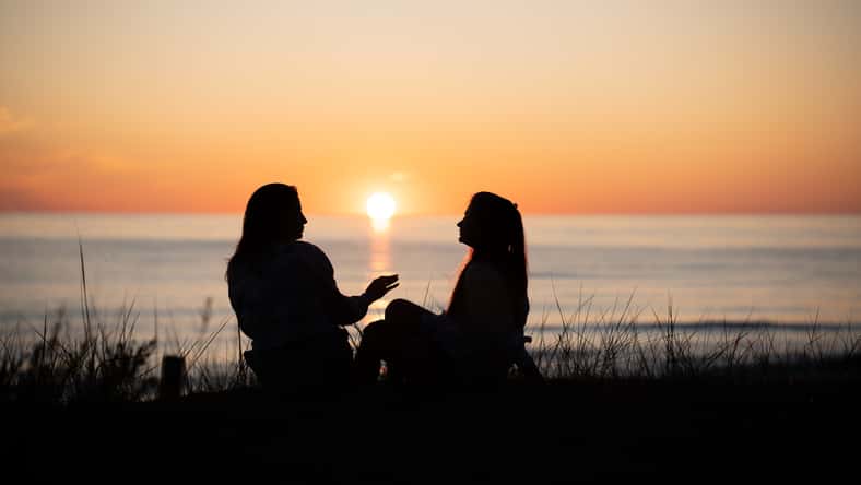 Silhouette of two female friends enjoying a conversation while sitting and watching the sunset on a sea beach. Back view