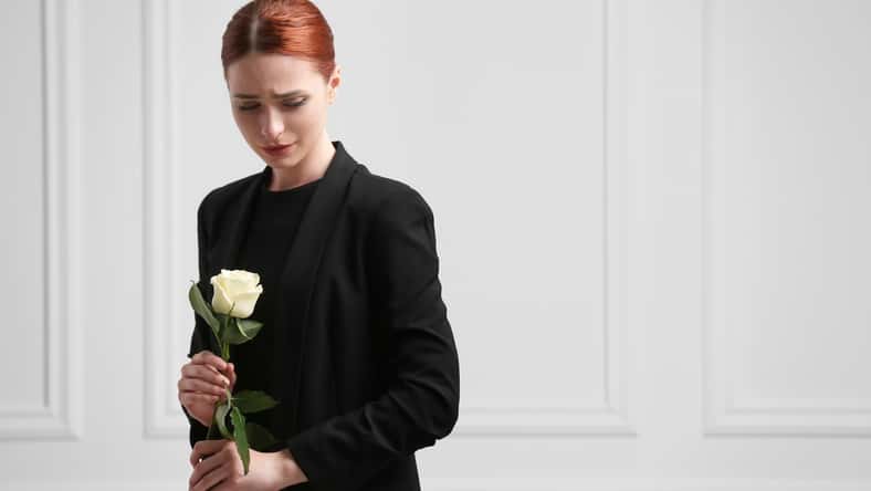 Sad woman with rose flower mourning near white wall, space for text. Funeral ceremony