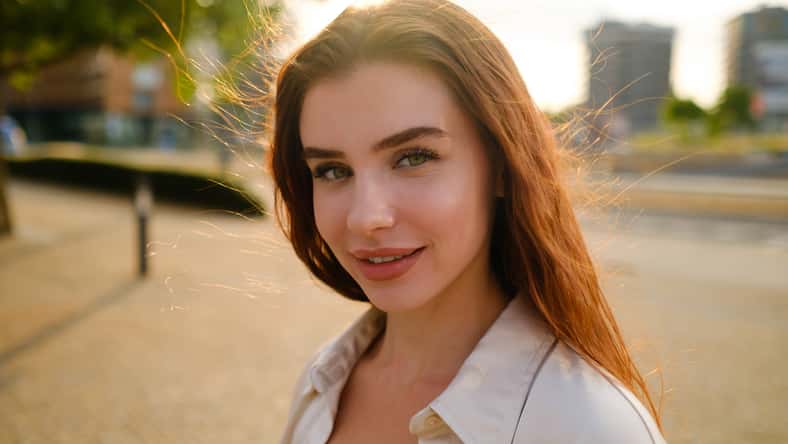 A portrait of a red-haired woman stands on the street, her vibrant locks framing her face.