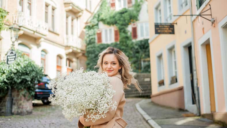 Portrait of beautiful young blonde woman with bouquet of white flowers in the center of European city. Happy model in casual spring clothes.