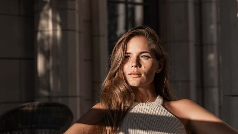 A woman with long brown hair sits in a chair, bathed in sunlight streaming through a window. She looks directly at the camera with a serious expression.