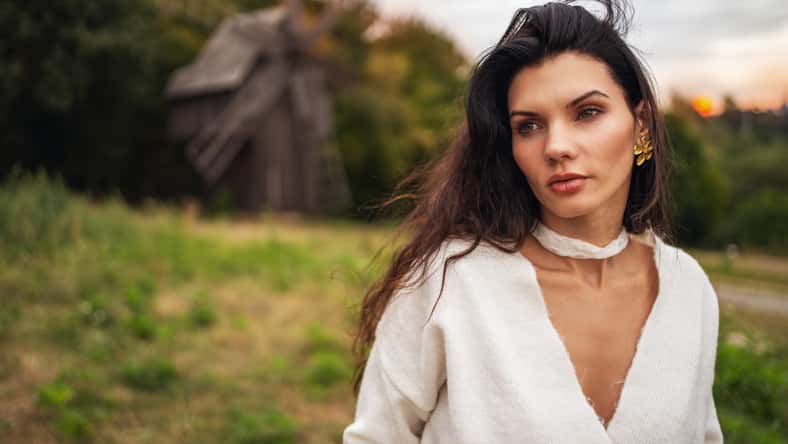 Retro style. Portrait of a young beautiful girl with black chic hair in a white dress on the background of a forest, a mill and a red sunset.