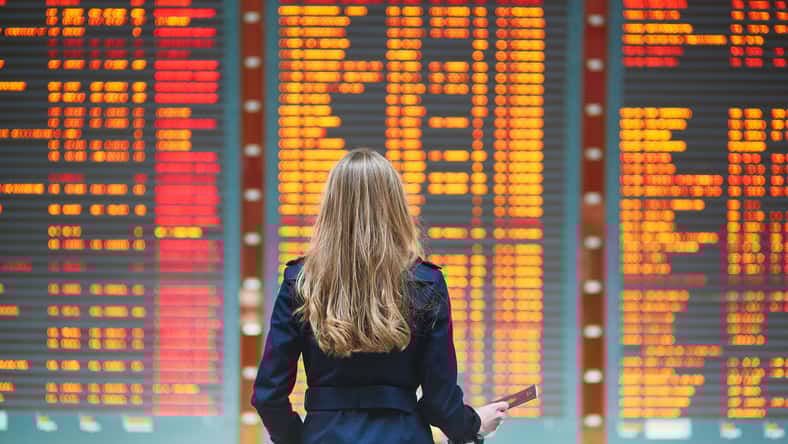 Young woman in international airport looking at the flight information board, holding passport in her hand, checking her flight