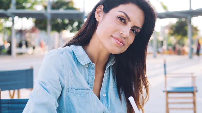 Beautiful caucasian female in casual wear enjoying free time on cafe terrace writing in notepad, charming young 30s woman journalist making notes for creating article blog on sunny day outdoors